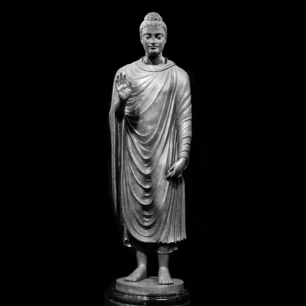 Standing Buddha statue with right hand raised in a gesture of reassurance, carved in stone against a dark background.
