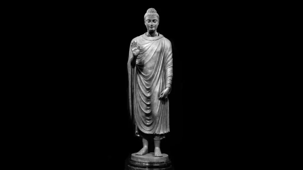 Standing Buddha statue with right hand raised in a gesture of reassurance, carved in stone against a dark background.